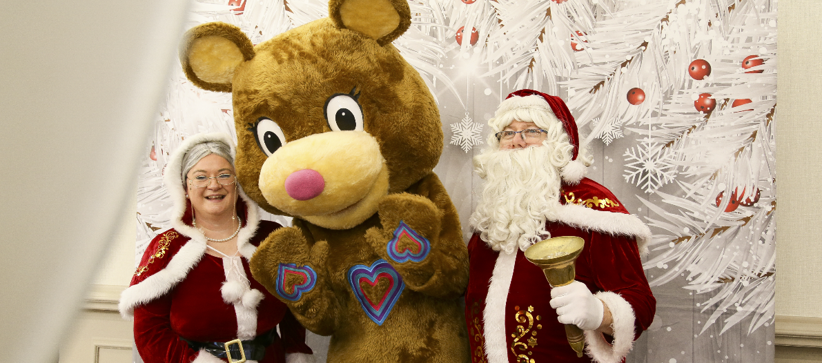 Charity mascot Katie Bear posing for a photo with Mr and Mrs Claus. White christmas background, santa is holding a bell