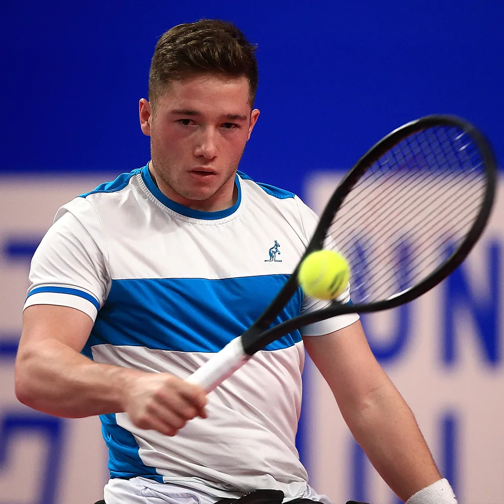 Alfie playing tennis, wearing a white and blue tshirt. He is mid-swing.