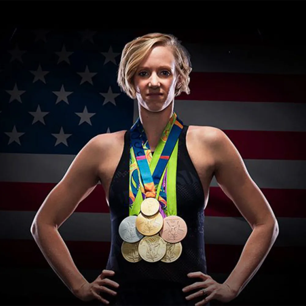 Dana is standing in a swimsuit with her hands on her hips, wearing 7 medals around her neck. The phot is dark, and she is lit from above creating a moody atmosphere. There is an american flag in the background. She looks strong.