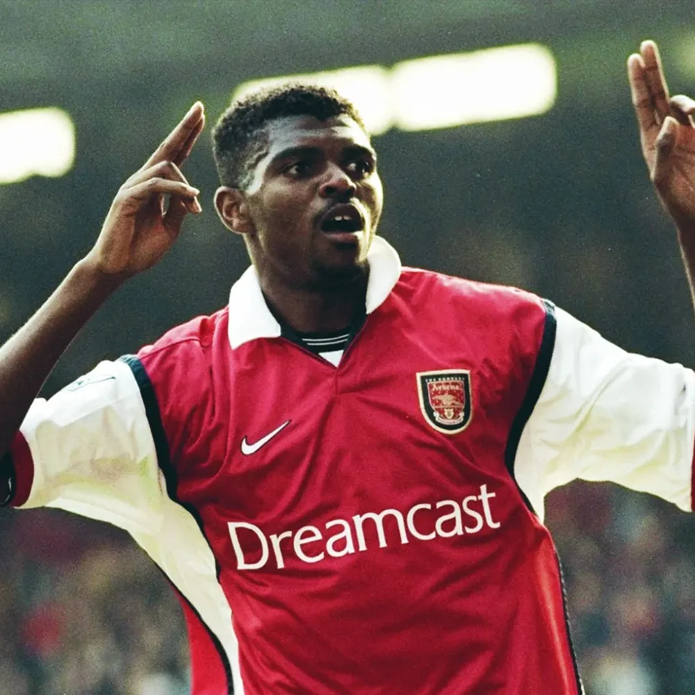 Nwankwo playing for Arsenal in the 90s. He is looking at the crowd after scoring a goal, with both his arms raised. He is wearing the Dreamcast Arsenal shirt in red.