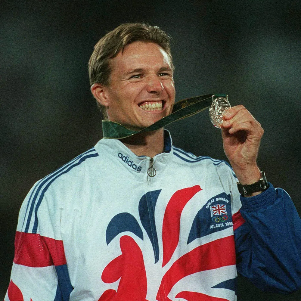 Roger is grinning. He is wearing a Team GB tracksuit and is holding up a medal after winning at the olympics in Atlanta