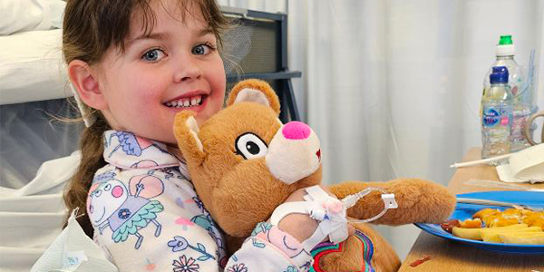 Girl in a hospital bed smiling and holding a Katie Bear teddy.