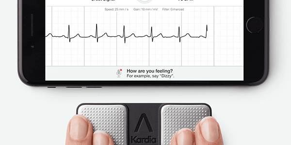 Fingers resting on a Kardia device that links to a mobile phone and reads a person's heart rate. At the top of the image is a smartphone showing a heart trace line.
