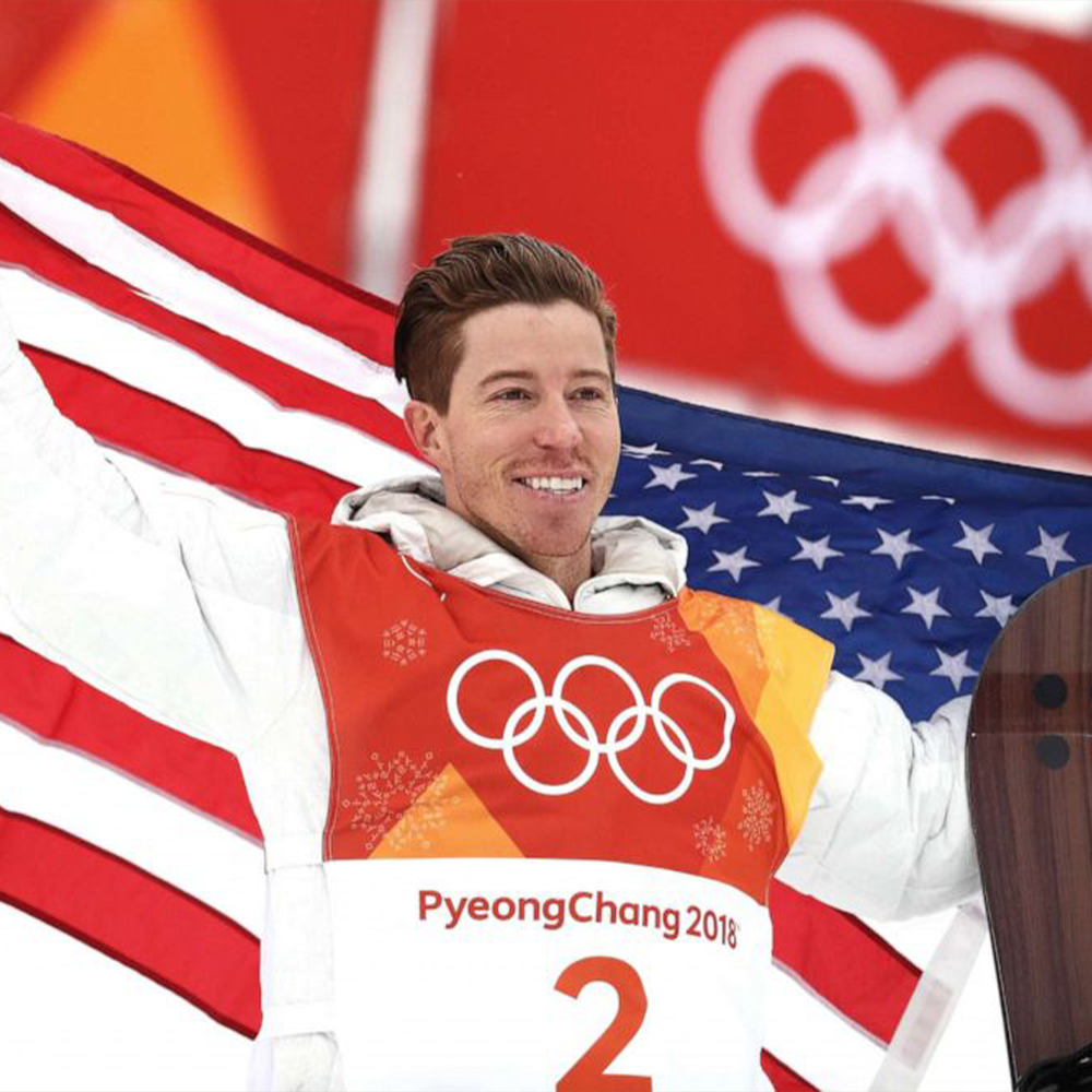 Shaun is smiling, holding a USA flag behind him and a snowboard is propped up in his left hand. He is on the podium about to receive an olympic medal. He is wearing an Olympics tabbard, PyeongChang 2018