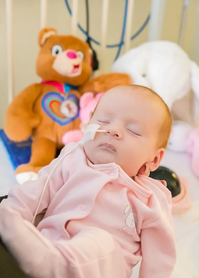Beau sleeping in her mum's arms in hospital. She has a feeding tube in her nose and is wearing pink. Her Katie Bear teddy is behind her on her bed.
