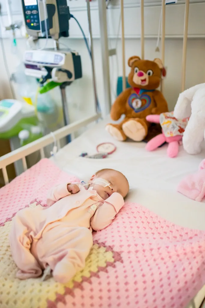 Beau in her bed in hospital, asleep wearing a pink baby grow and laying on a pink blanket. Kati eBear teddy is at the head of the bed with her.