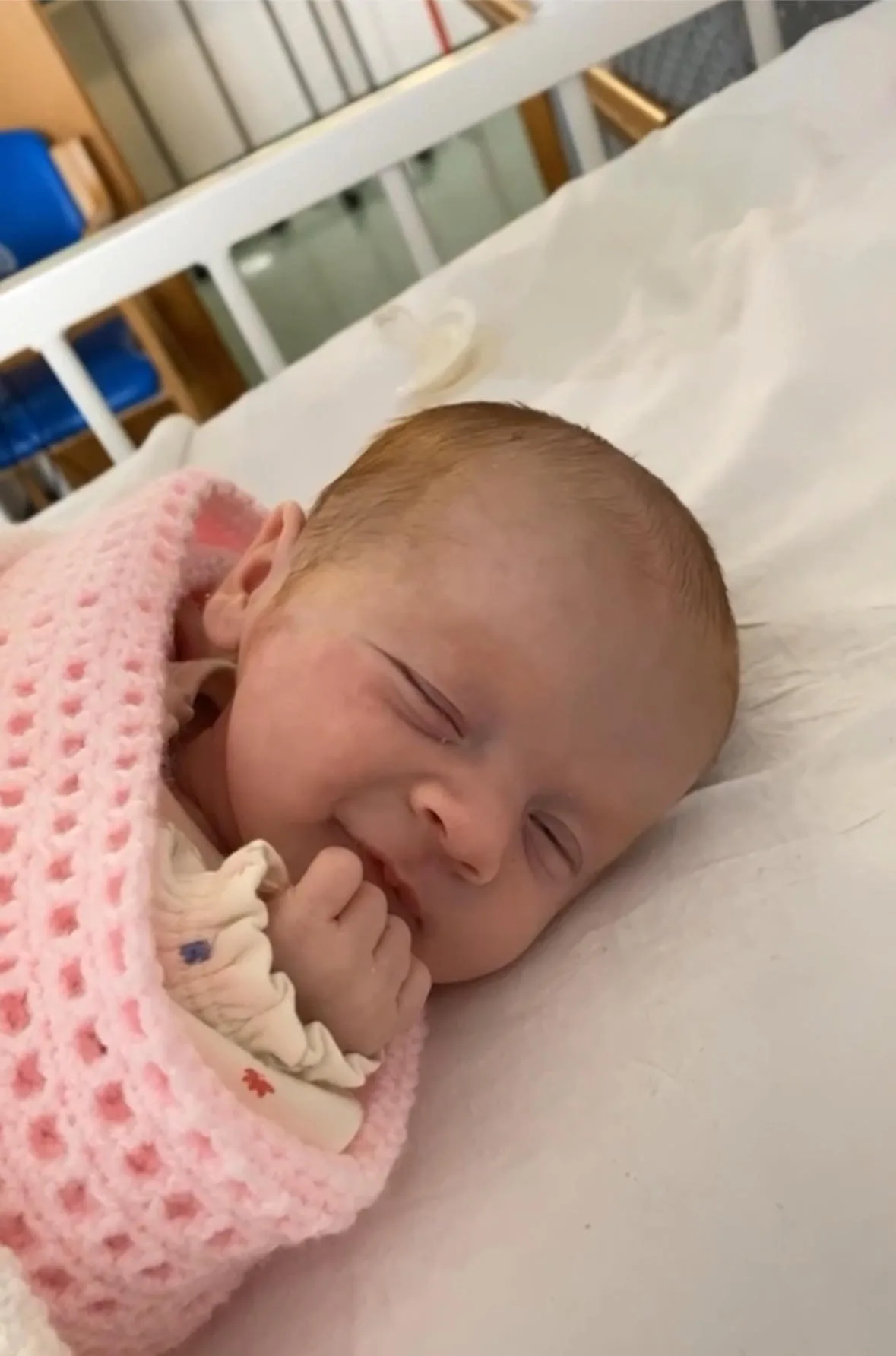 Baby Beau, asleep but smiling in her bed in hospital. She is covered in a pink blanket and wearing a pale yellow knitted jumper.