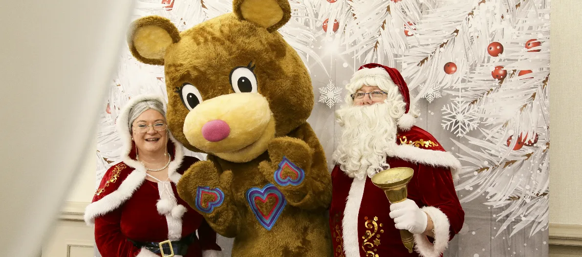 Charity mascot Katie Bear posing for a photo with Mr and Mrs Claus. White christmas background, santa is holding a bell