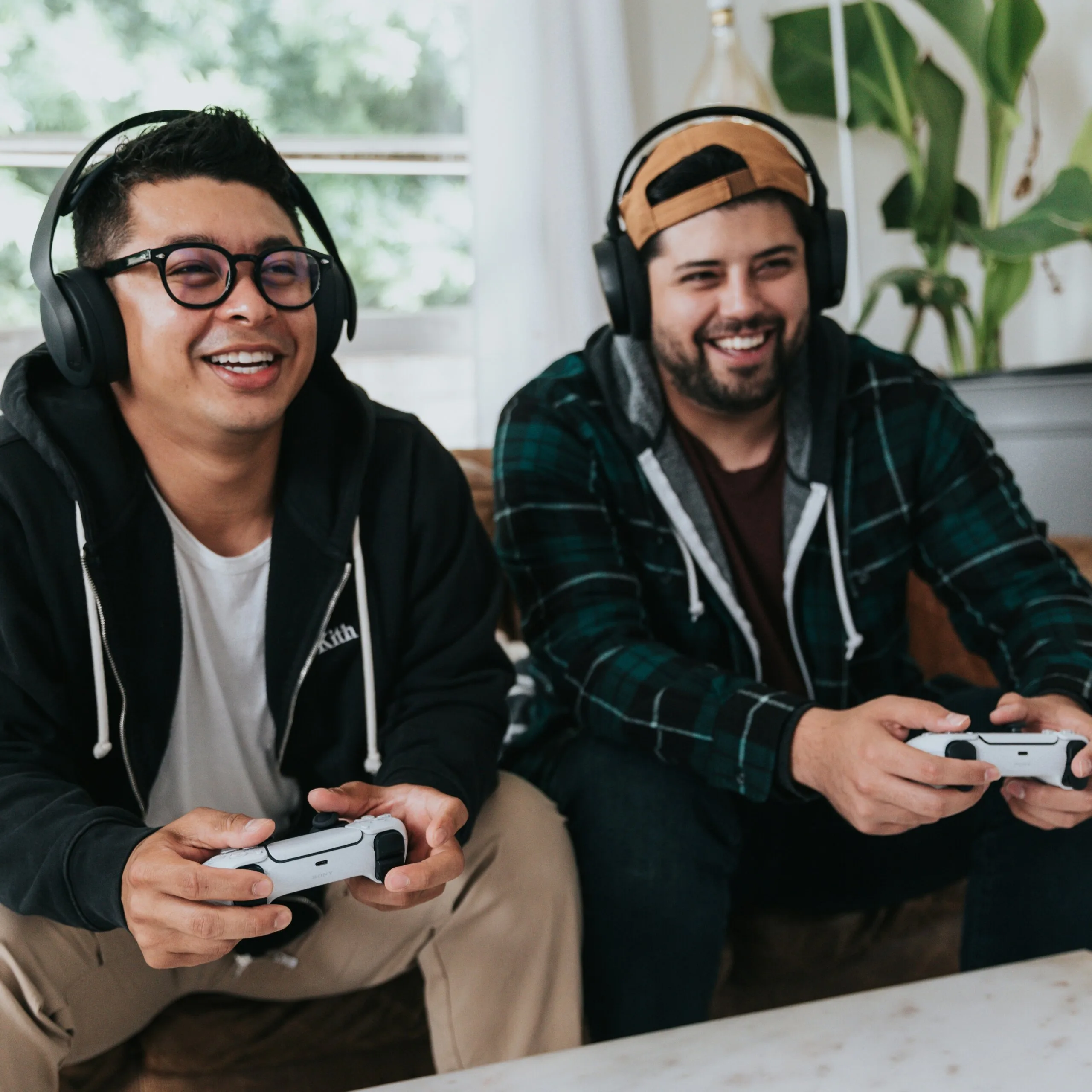 Two young men holding playstation controllers and wearing head phones  smiling while they are playing a game together. One is wearing glasses, the other is wearing a backwards snapback